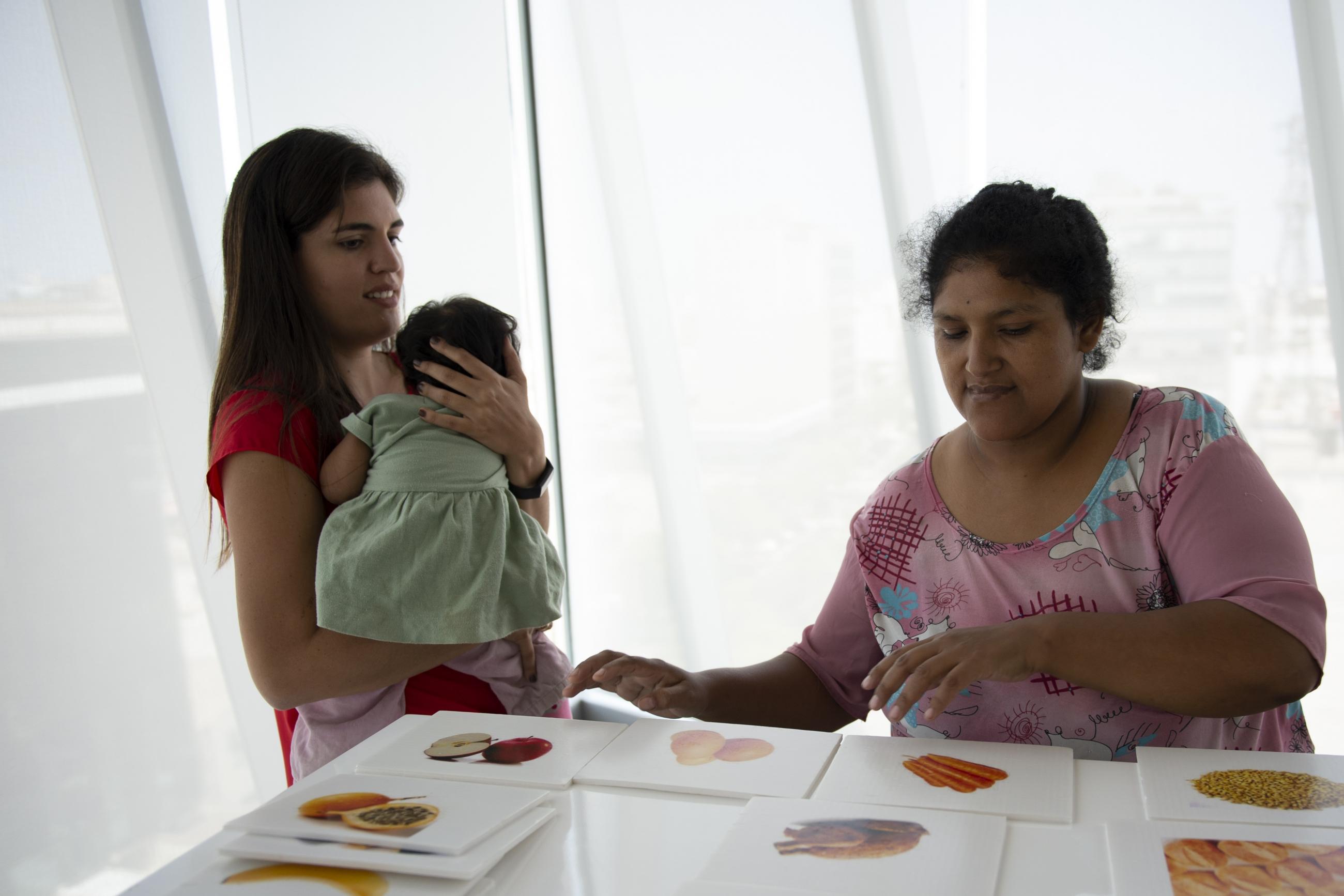 Two women stand at a table, looking at pictures of healthy food options. One woman is holding a baby.
