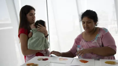 Two women stand at a table, looking at pictures of healthy food options. One woman is holding a baby.